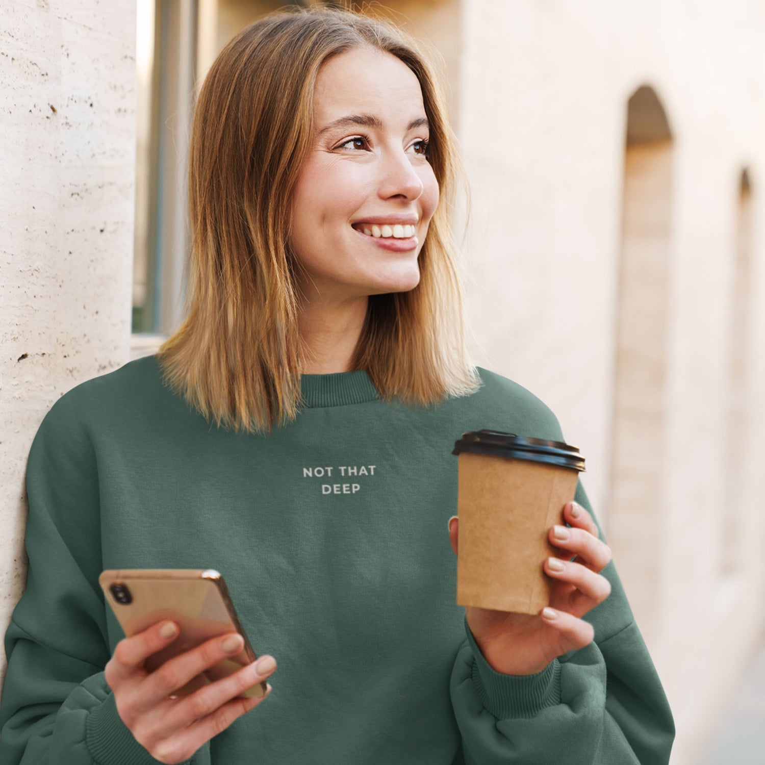 Woman wearing a green sweatshirt with 'Not That Deep' text, holding a phone and a coffee cup.