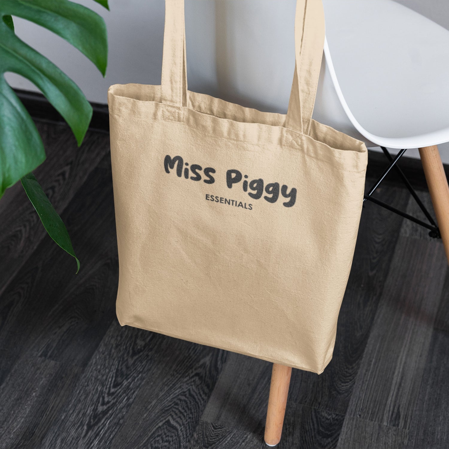 Beige tote bag with 'Miss Piggy Essentials' text on a wooden floor.