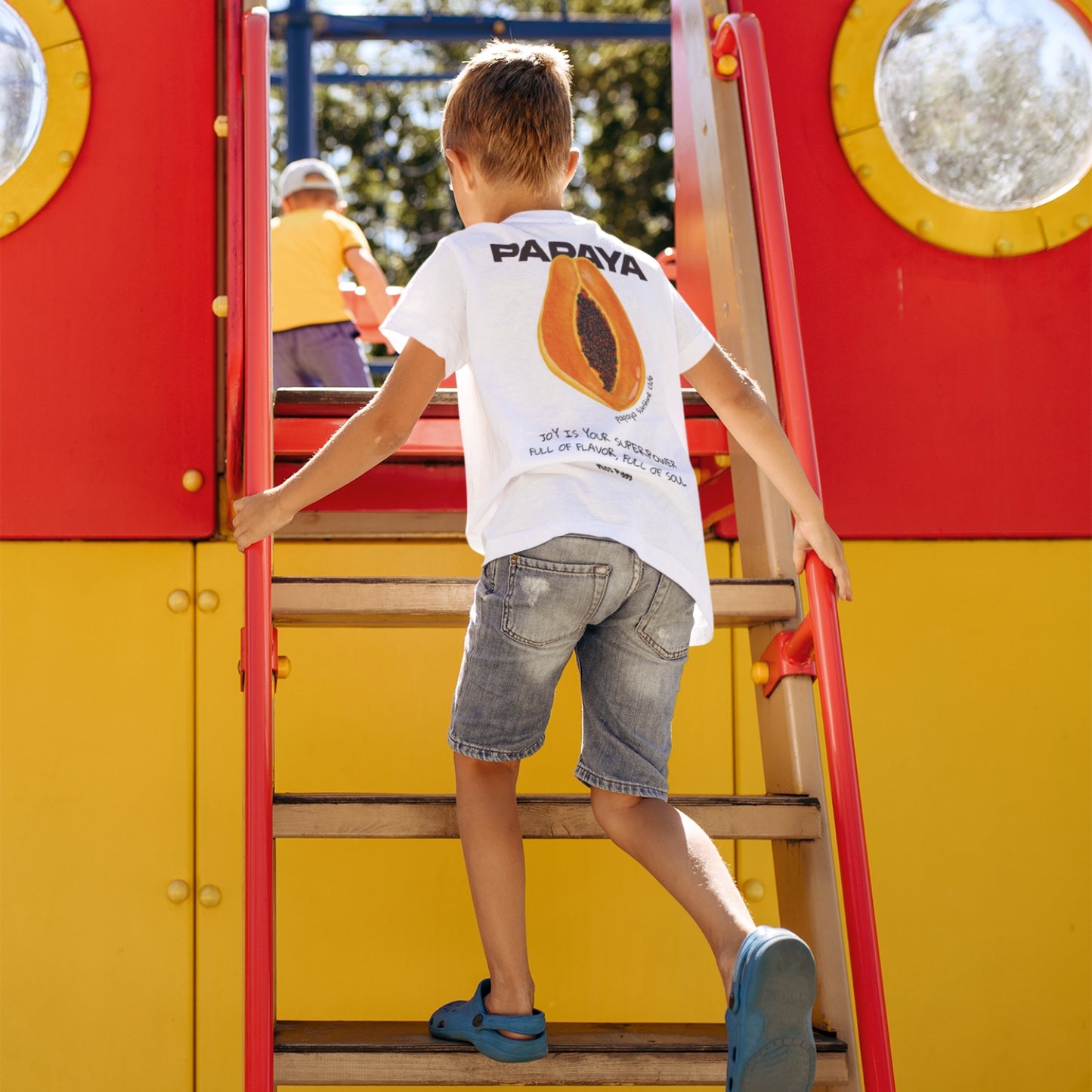 Child wearing the PAPAYA t-shirt in white. Unisex organic cotton design with soft texture and playful energy, shown in a light and joyful setting.