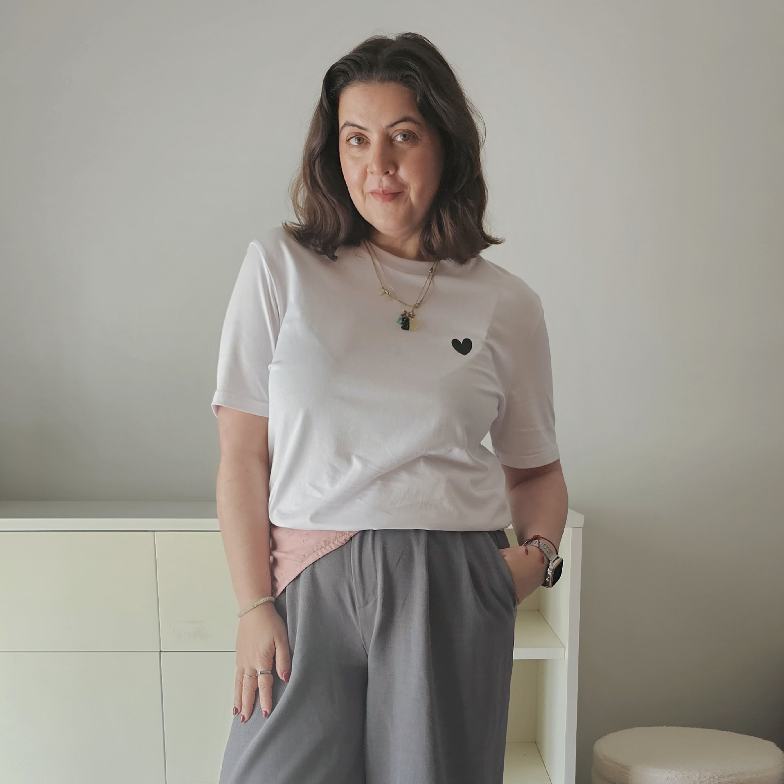Rita Costa wearing a white t-shirt with a black heart design and gray pants standing in a room.