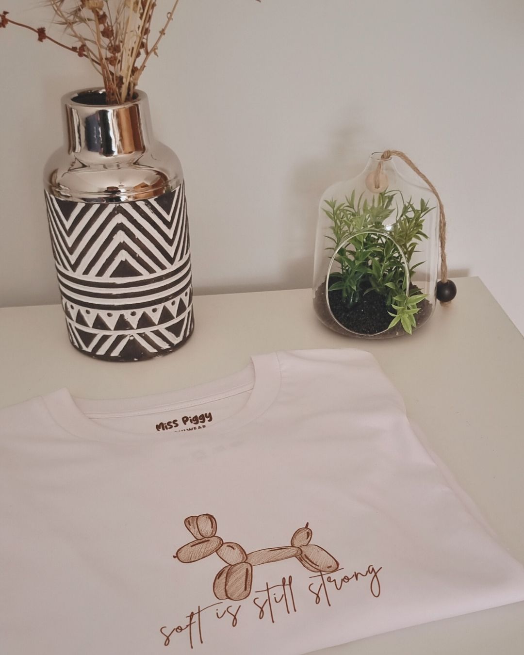 White t-shirt with balloon dog design and text on a white surface with decorative items.